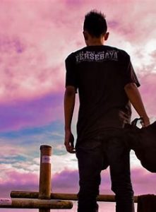 A man looking back. He's looking towards a sunset, consisting of a cloudy sky with pink tinges and a streak of blue and purple across it.