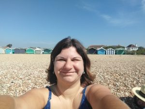 A selfie of me taken on a pebbled beach with beach huts along the horizon and a blue sky.