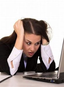 A lady with her head low down with her hands eitherside of her head. On one side she has her hair wrapped around her hand. She is looking at the laptop in front of her